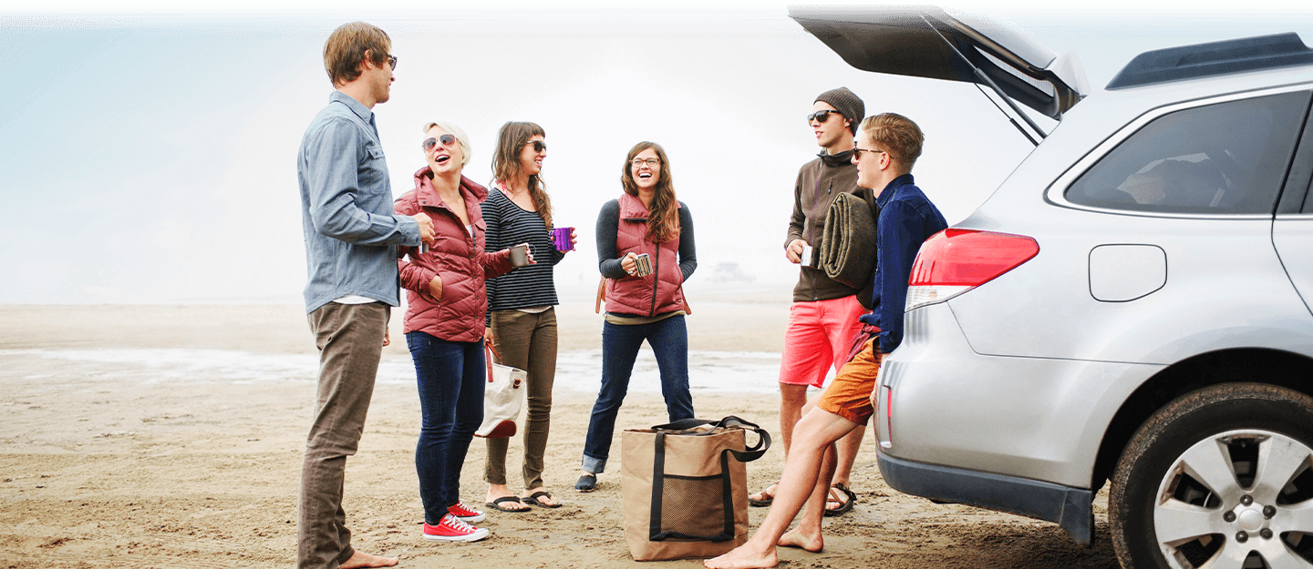 Friends hanging out laughing at the beach by their silver Subaru station wagon 