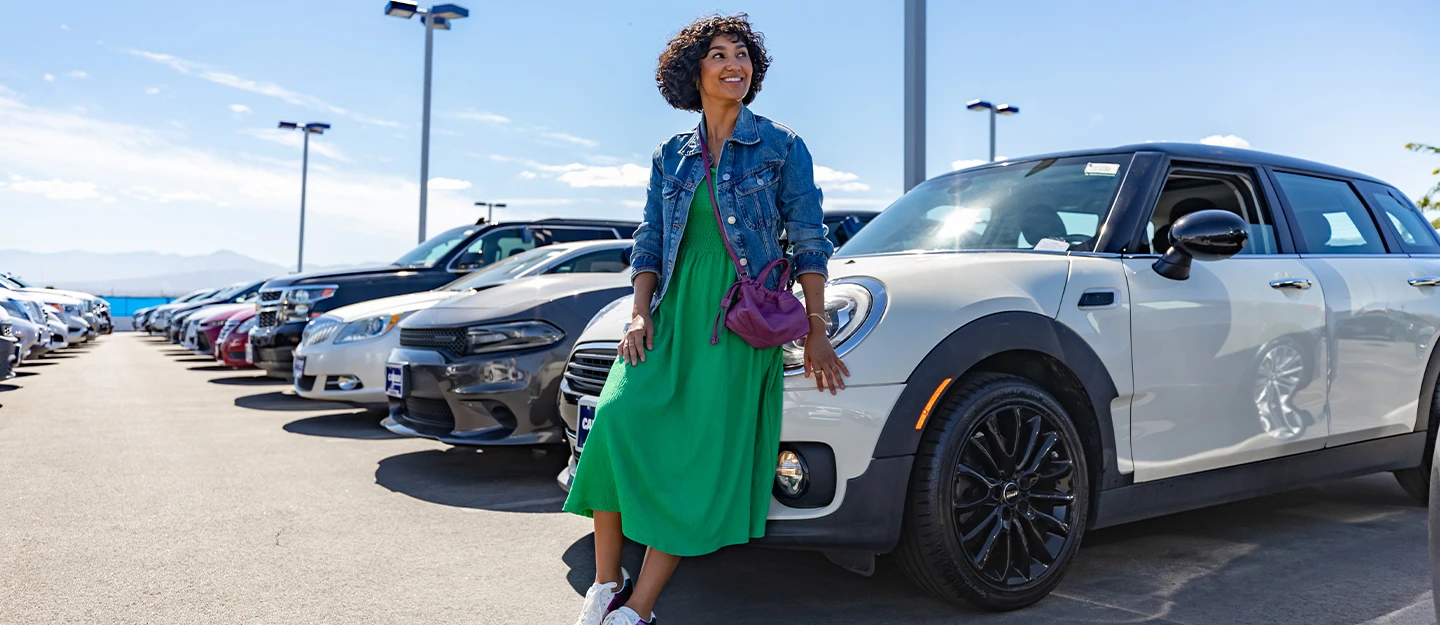 Woman in green dress leaning on white Mini Cooper as she walks the lot at CarMax in search of a new car the sun is shining and mountains are in the distance 
