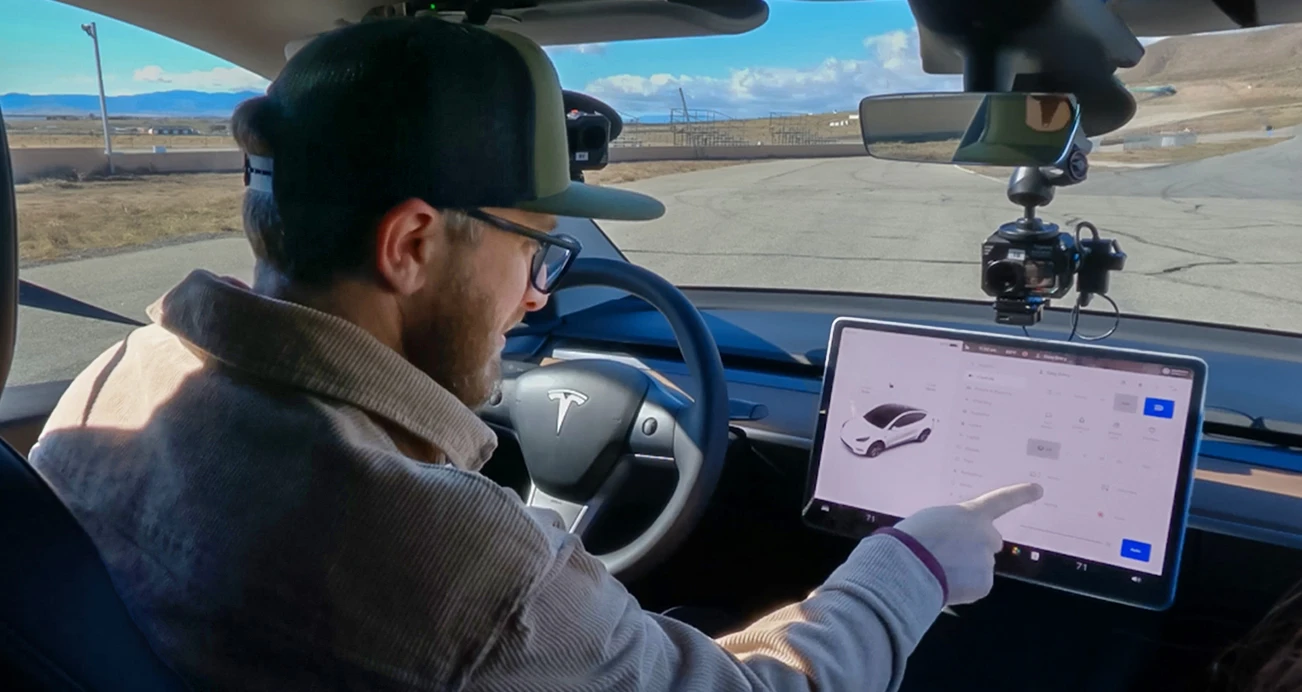 Man wearing green baseball cap, beige jacket, and black glasses, selects option on touch screen from the drivers seat of a Tesla.