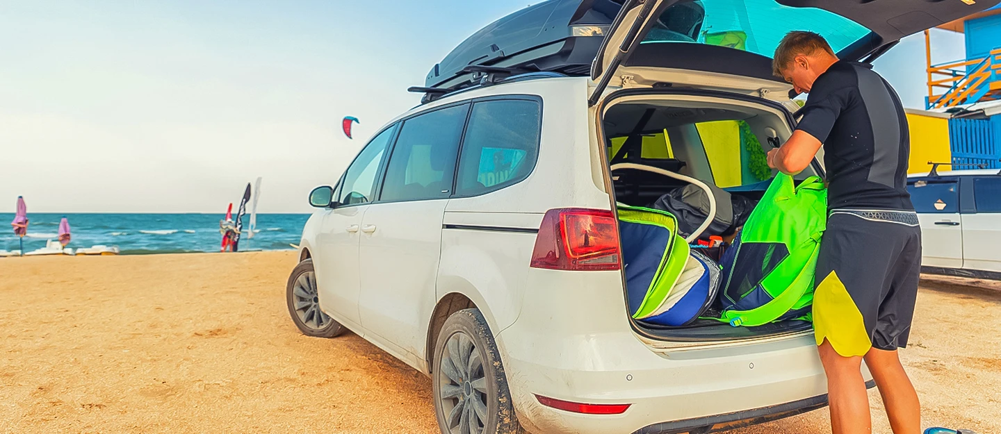 Man unloading beach gear from a white minivan parked directly on a sandy beach, preparing for a day by the ocean.