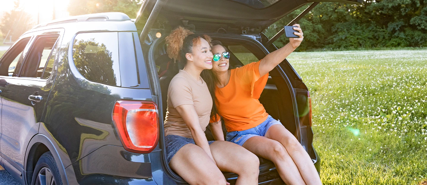 Two friends sitting with the trunk open of their large SUV they are smiling and taking a selfie together the SUV is parked in a field of white flowers 