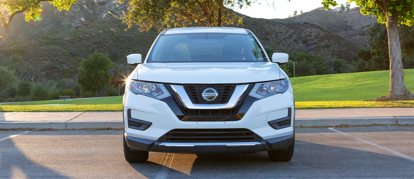 White Nissan Rogue parked in front of rolling green hills; a tree sits in the right side of the frame