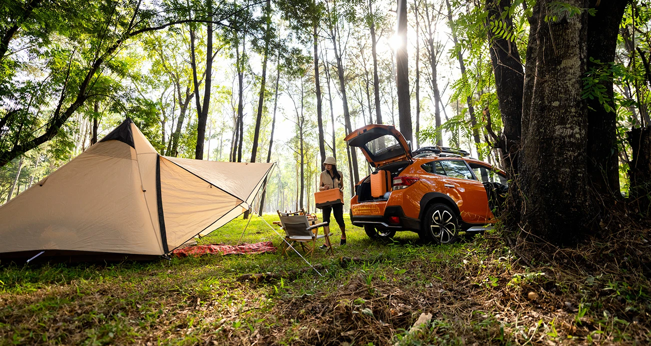 Woman setting up campsite in woods pulling supplies from her orange Subaru Outback SUV parked next to tent