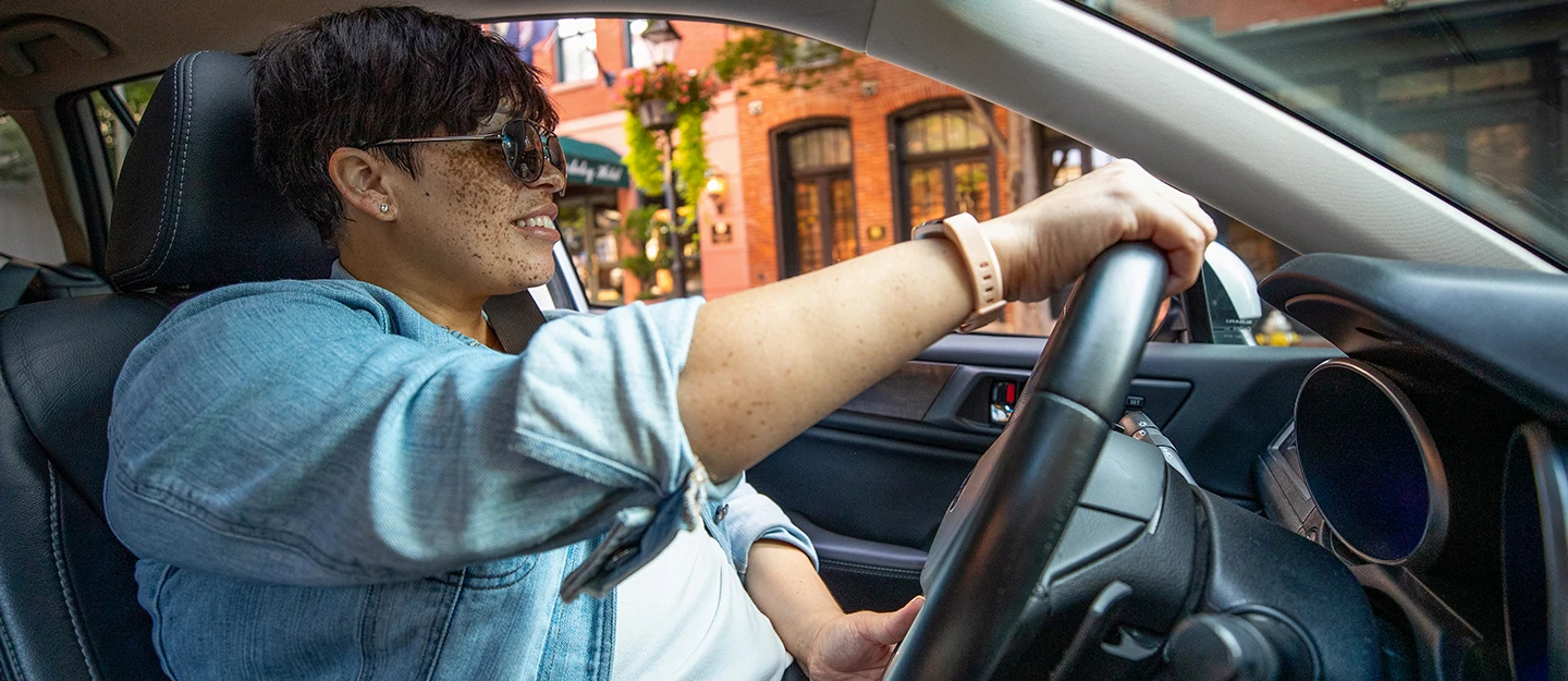 Lady in denim jacket and sunglasses smiling while sitting behind the wheel of her car cruising a city street