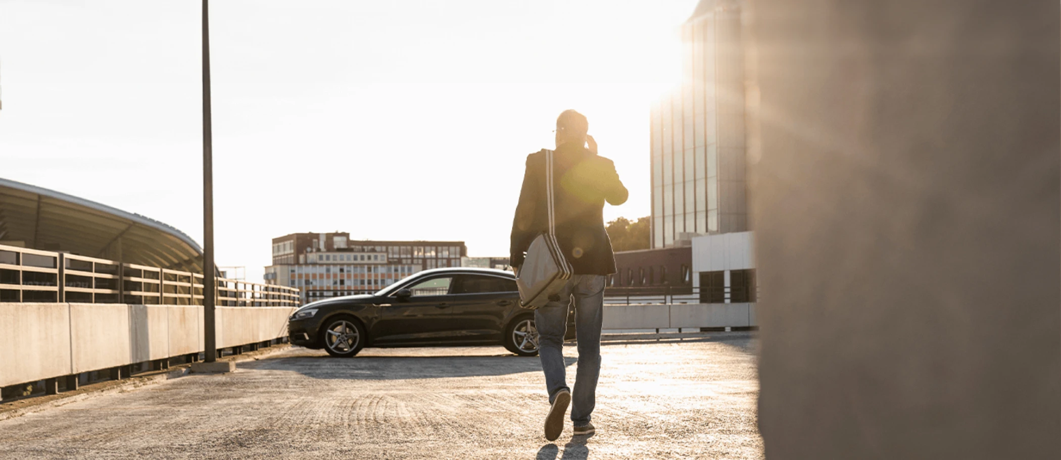 Man talking on his phone holding a laptop bag walking towards his sedan parked on roof of parking deck as the sun sets