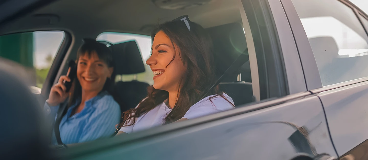 Two women sit in a compact car, laughing together. The passenger is also using her phone, capturing a candid moment of joy.