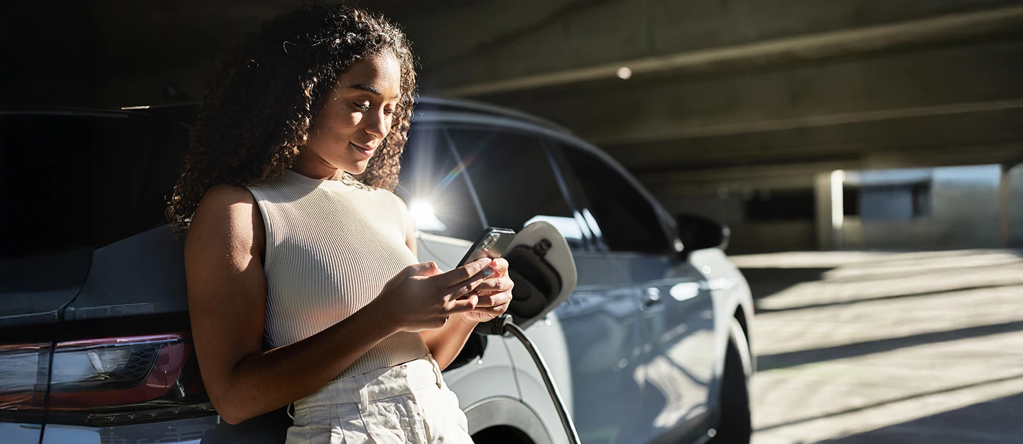 Woman leaning on her electric SUV while it charges. She is smiling while looking at her phone