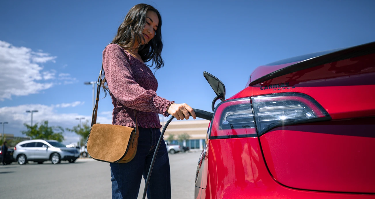 Woman in long sleeved top with a purse on her shoulder plugs charger into red Tesla in parking lot.