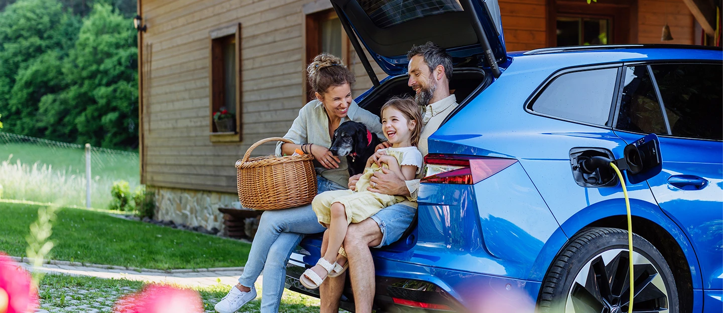 A family relaxes in the back of a hybrid SUV while it charges. The mother, holding a picnic basket, smiles at her daughter who sits on the father's knee. Their older dog rests contentedly between them.