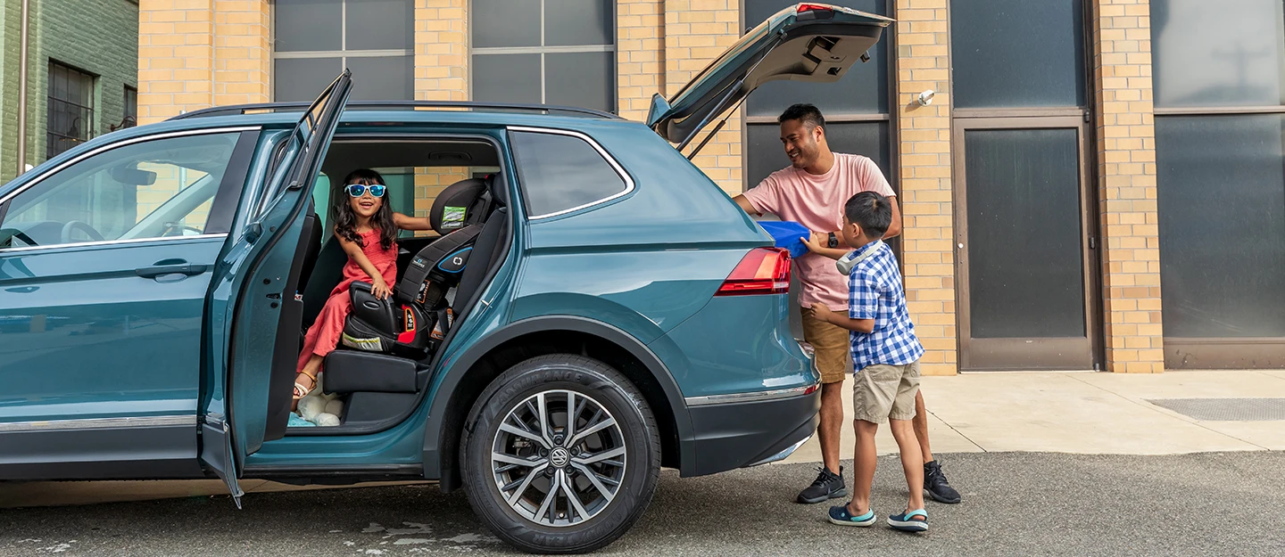 Father and son unload a bin from the back of a blue Volkswagen SUV while daughter wearing sunglasses smiles in the backseat with the door open