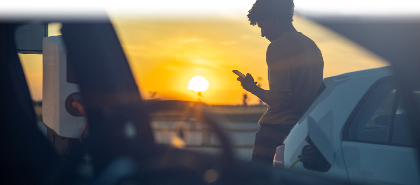 Person leaning on electric vehicle as it charges staring at phone sun is setting in background