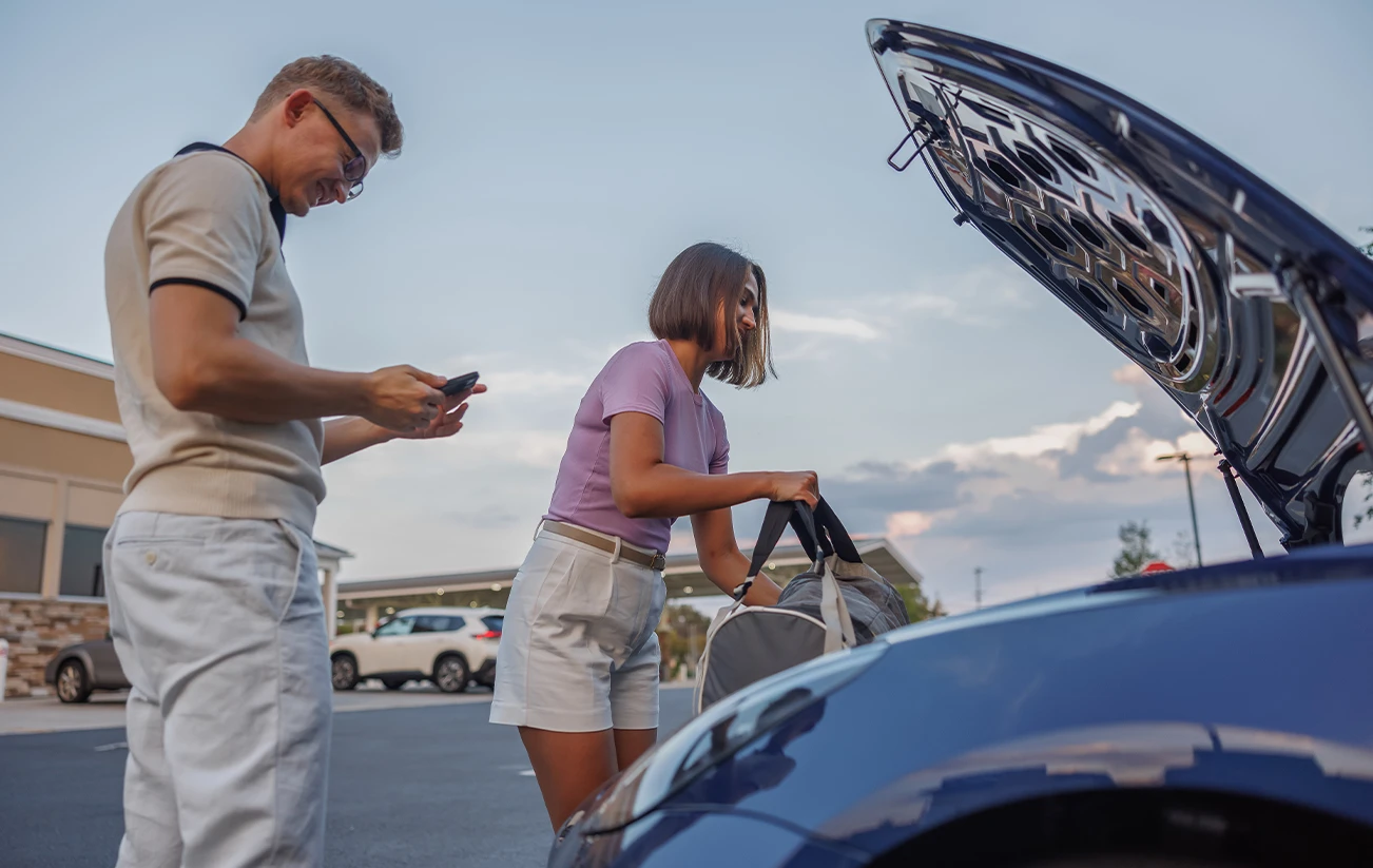 Couple standing by electric vehicle woman packing overnight bag into vehicles frunk while the man is checking his phone 