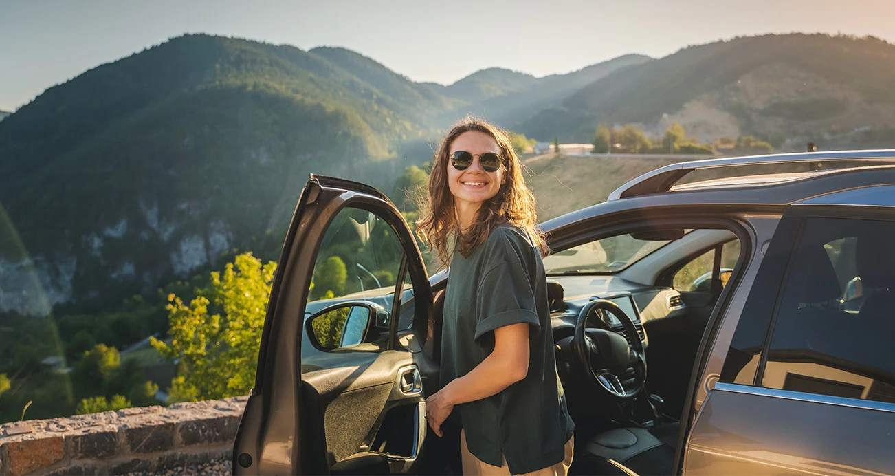 A woman smiling wearing sunglasses steps out of her car at a breathtaking mountain lookout. 