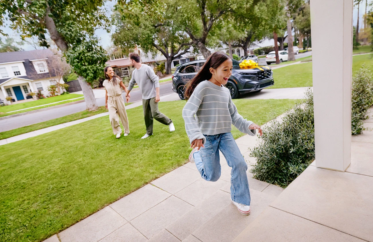 Family smiling in their front yard as their new to them CarMax car is delivered to their driveway complete with yellow bow on hood of car