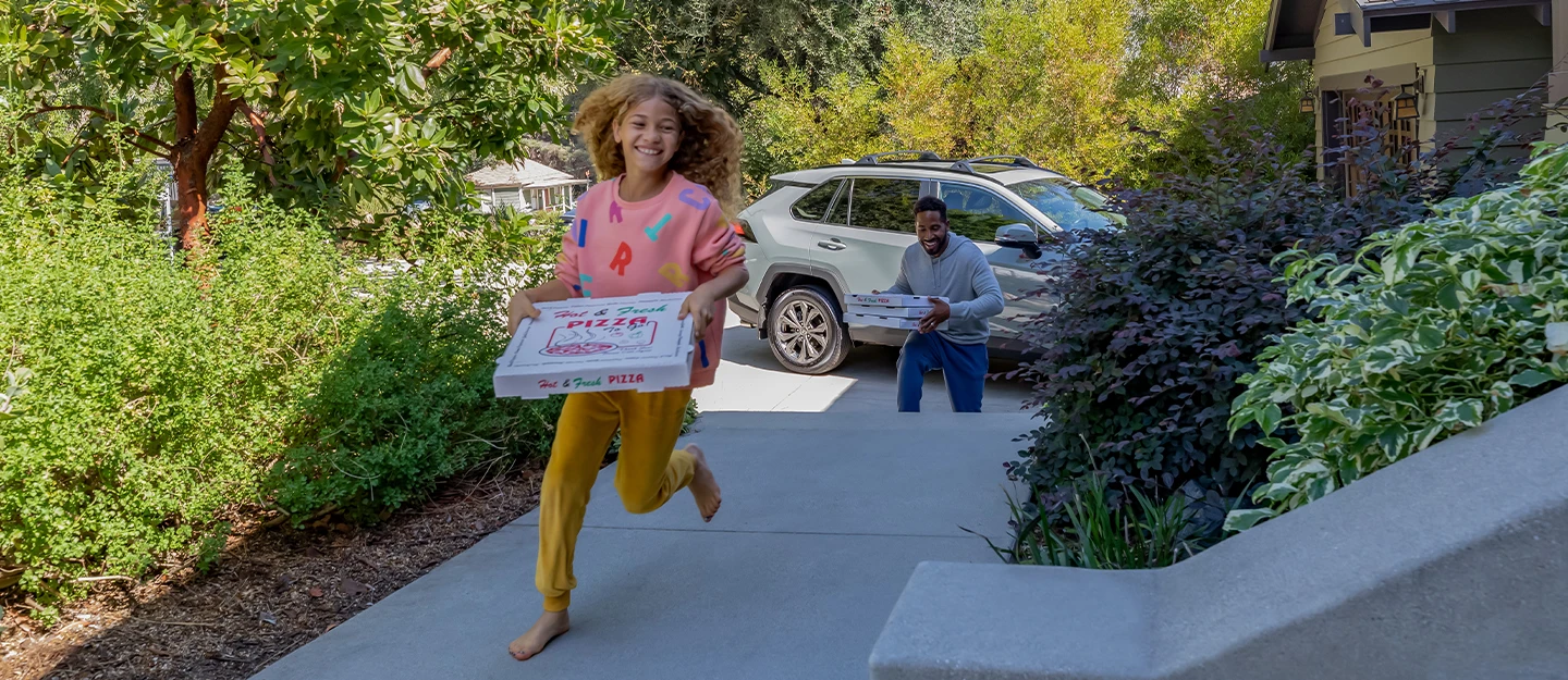 Daughter running up steps to home with a pizza box in hand father follows behind holding 2 boxes Toyota RAV4 parked in driveway