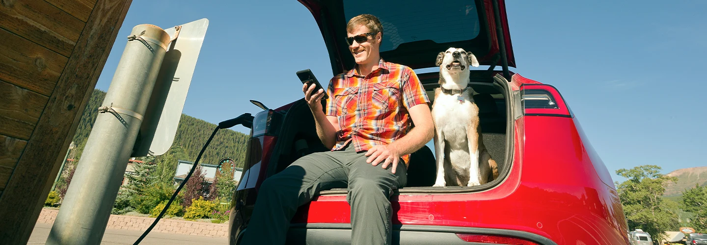 Man sitting with his dog in back cargo space of a red electric car playing on his phone while car charges