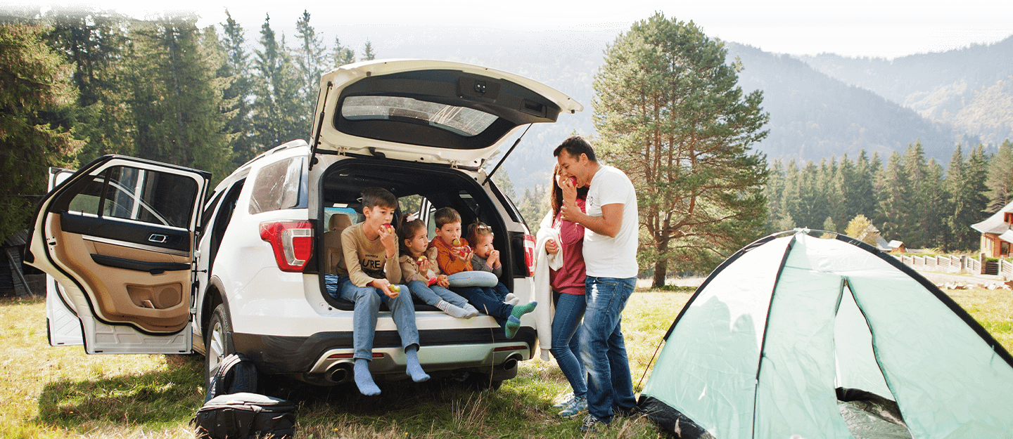 Family sitting in open trunk of white SUV next to camping site