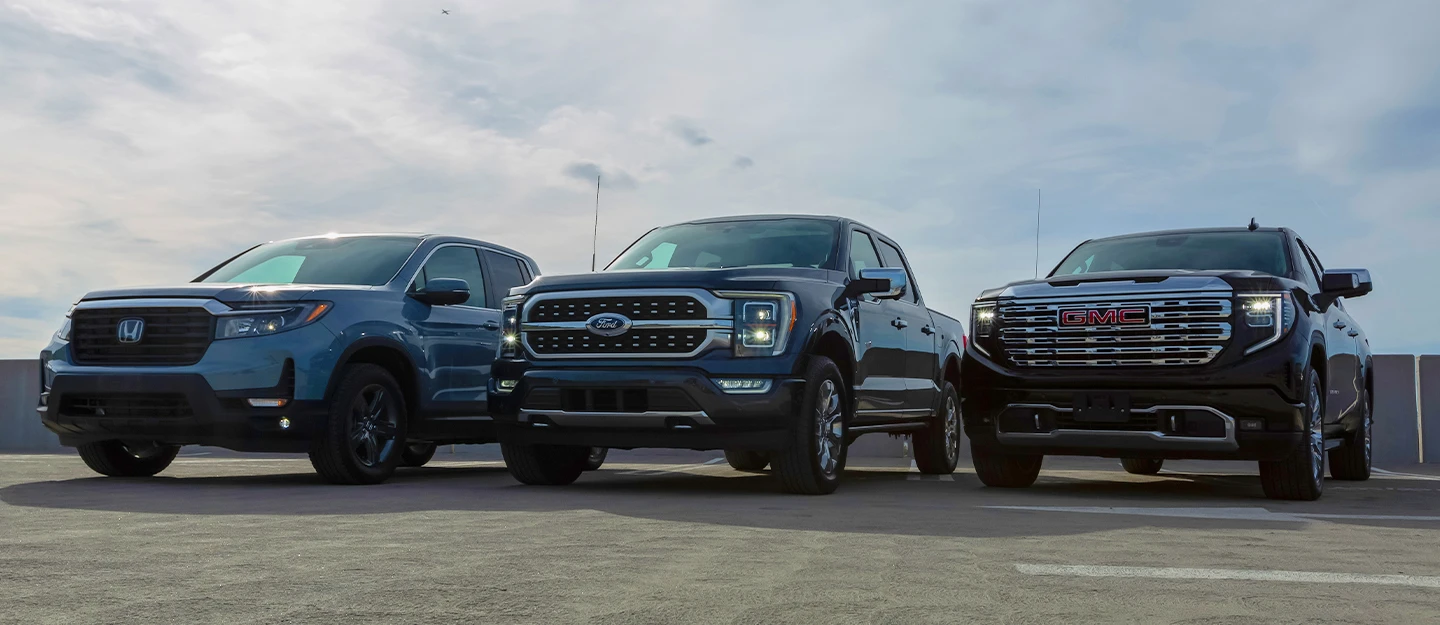 Three full-size pick up trucks: Honda Ridgeline, Ford F150 and the GMC Sierra parked side by side on the top level of parking garage. Blue skys above