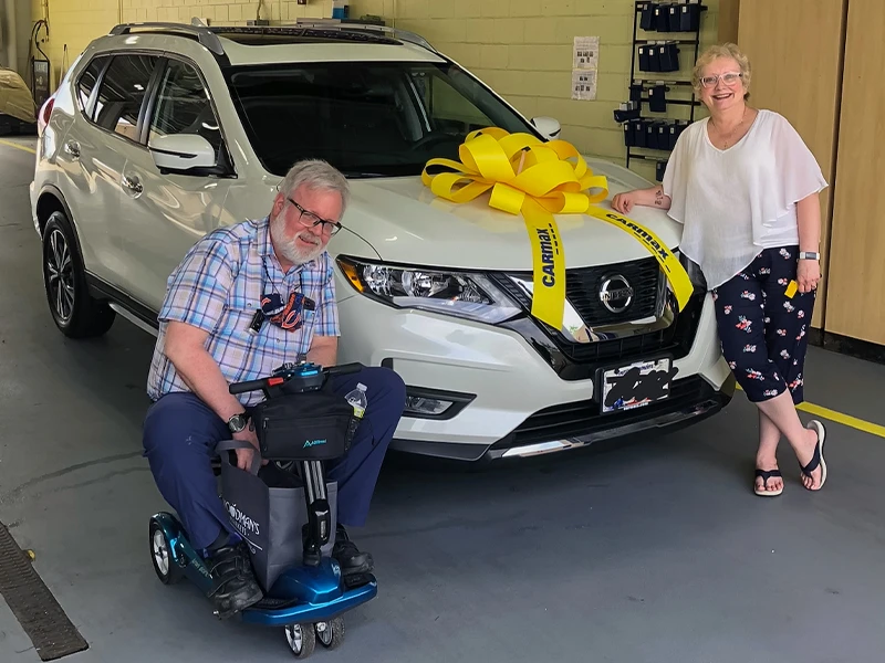 CarMax customers Cathy and Ken at the purchase of their 2020 white Nissan Rogue. Cathy leaning on vehicle with yellow bow on hood, Ken on his mobility scooter both smiling 