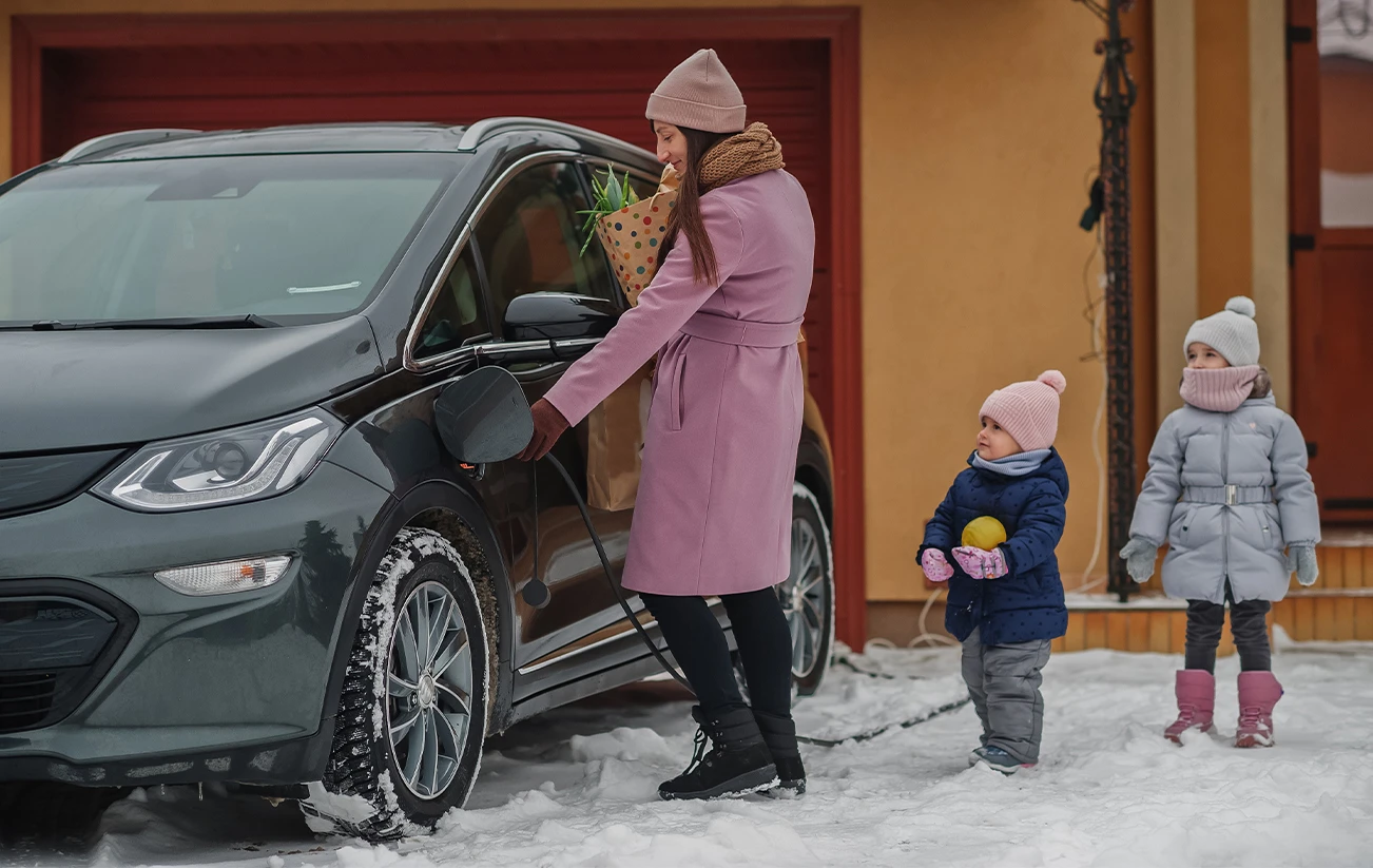 Electric vehicle parked in snow covered driveway woman unplugs ev with one hand and carrys groceries in the other two small children follow 