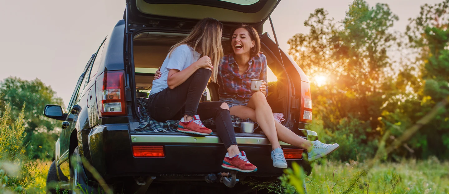 Two girl friends sitting with the trunk of their SUV open as they sip coffee laughing 