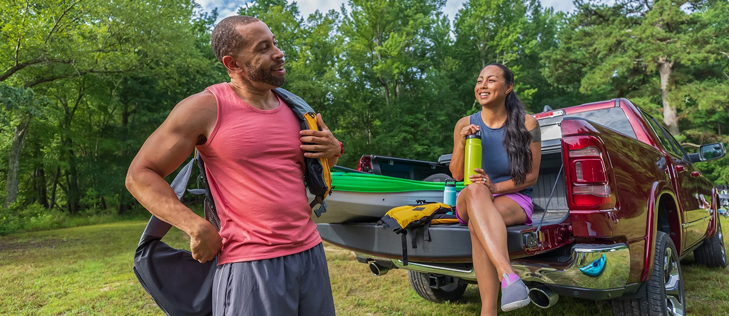 A woman sits in the back of a red pickup truck with a kayak placed next to her. A man is unloading a backpack from the truck and appears to be talking to her. The truck is parked in a serene, wooded area