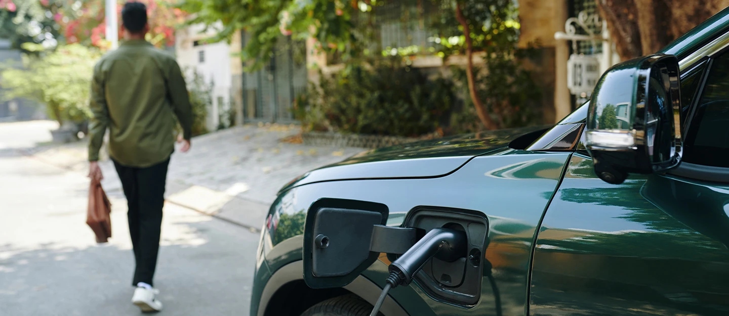 Electric vehicle plugged into charger in foreground, driver walks away from camera with a bag in hand.