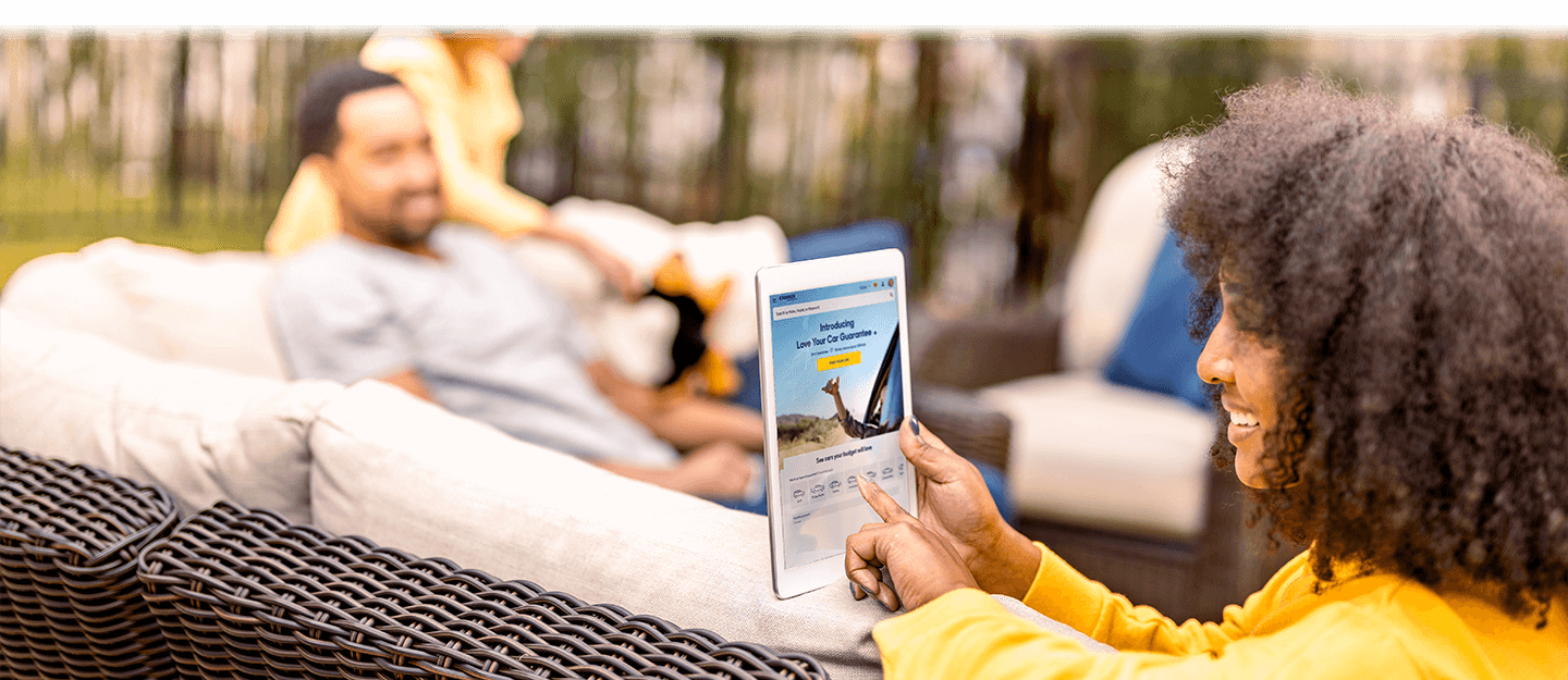 Woman holding ipad on CarMax website sitting in backyard on outdoor setting. Family in background. 