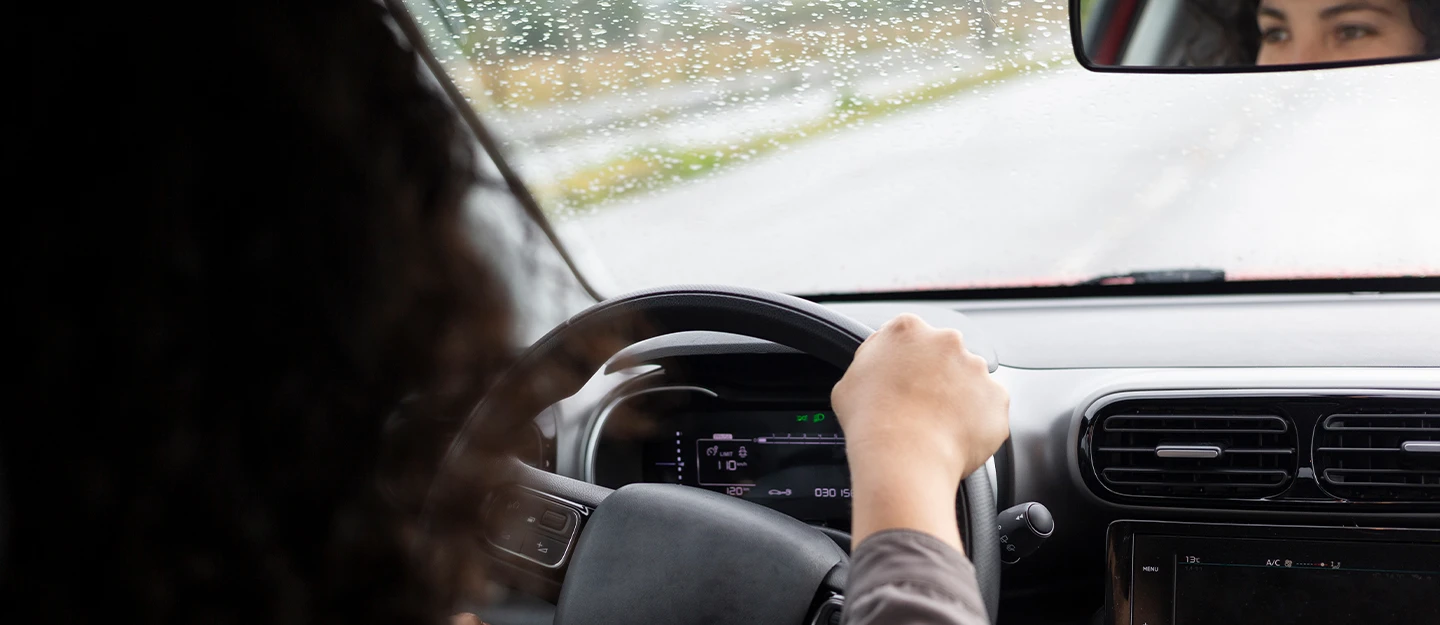 A woman with her hands on the steering wheel of an electric car, driving through rain with water droplets on the windshield.