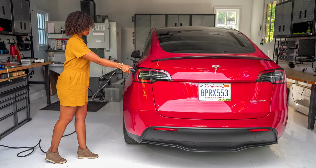 Woman in yellow dress plugs charger into red Tesla Model Y inside of a garage surrounded by tidy storage and tools.