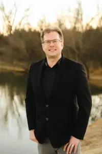 Author Andy Jensen wears a black blazer and shirt while standing outside smiling, with one hand in his pocket.