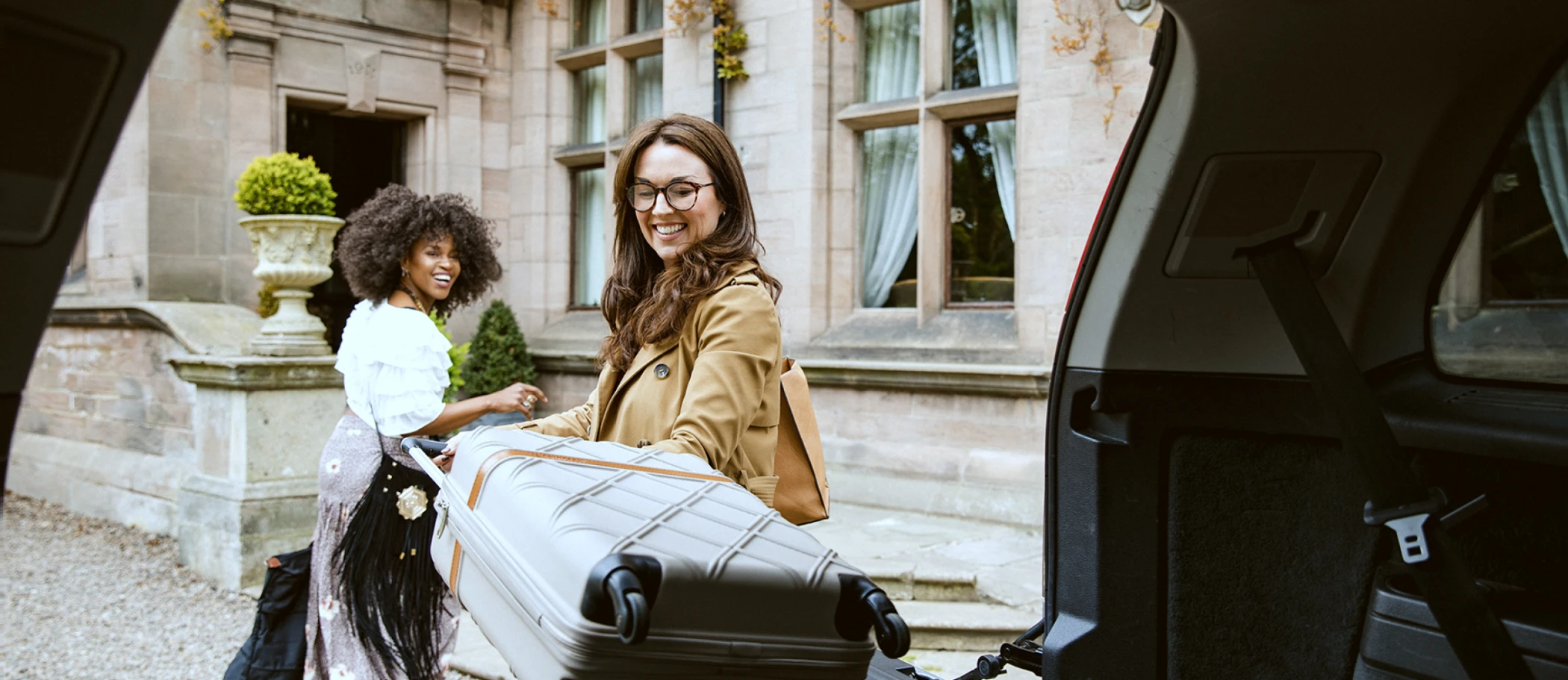 A woman is pulling her suitcase out of the back of a luxury SUV and another woman turns back to watch her