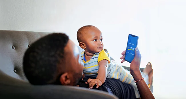 How to Sell Your Car: Man laying on couch with baby scrolling through CarMax website on his phone | CarMax