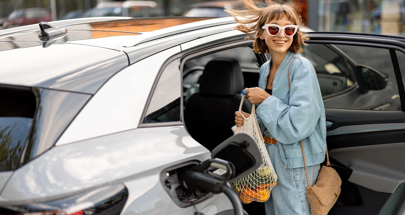 Smiling woman wearing sunglasses places groceries in the back seat of an electric vehicle while it charges.