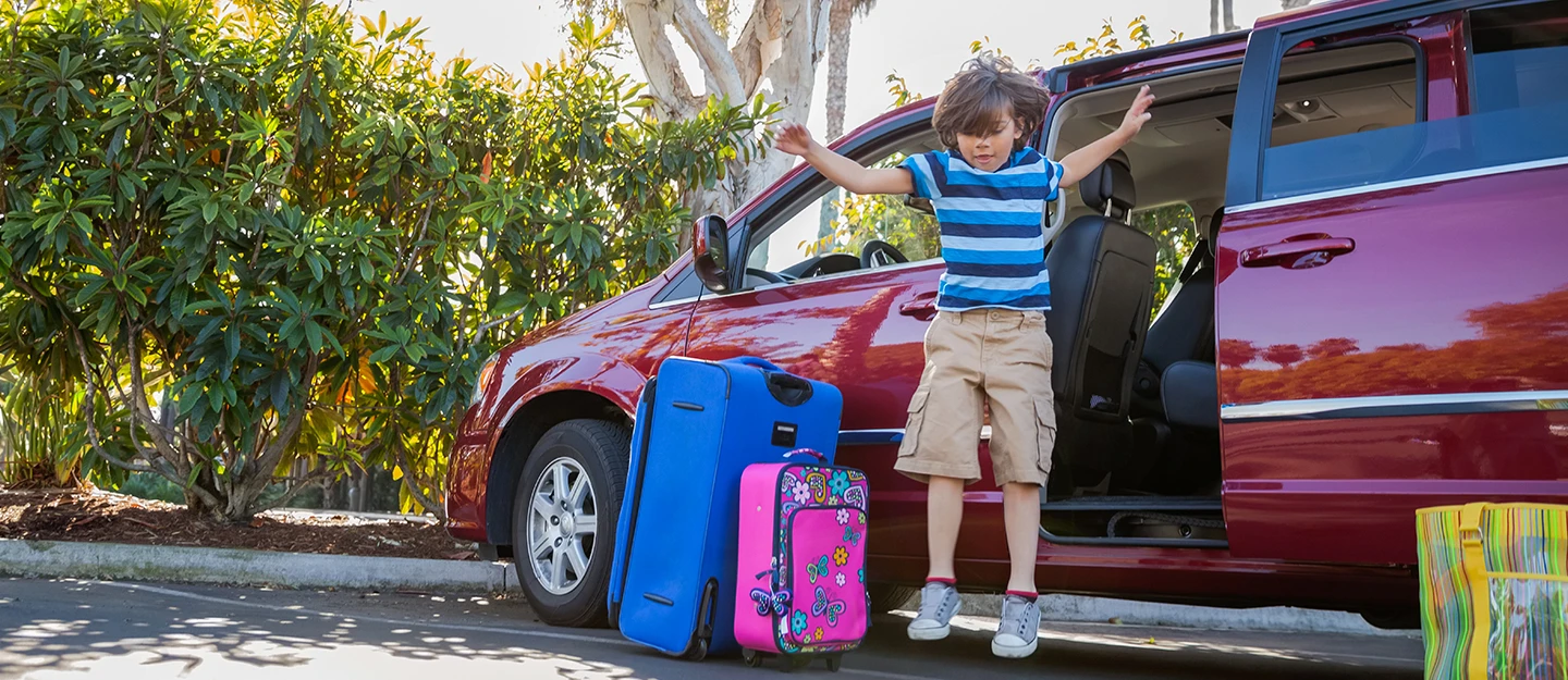 Young boy jumping out of parked minivan as luggage is unloaded 