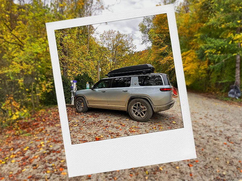 A Rivian sits plugged in and facing away from the viewer at an Electric Vehicle charging station with a black roof storage container on top of the roof racks.