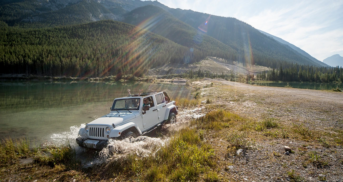 White Jeep Wrangler off roading along stream with mountains in the distance 
