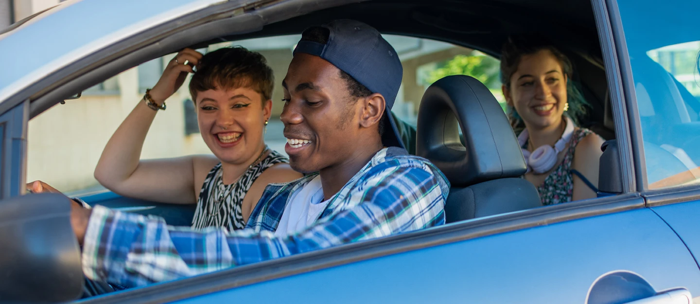 Three friends sit laughing, viewed through the open window of a blue subcompact car. The driver wears a plaid shirt and backwards blue baseball cap; the passenger wears a sleeveless zebra print top; the third friend sits in the back seat wearing a black and white blouse and pink headphones around her neck.