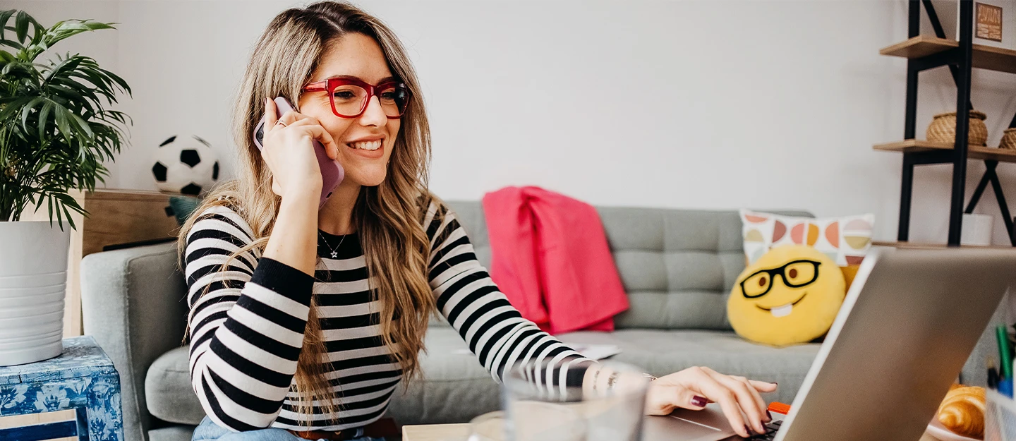 Woman sitting at a coffee table in her bedroom, speaking on the phone while working on her laptop.