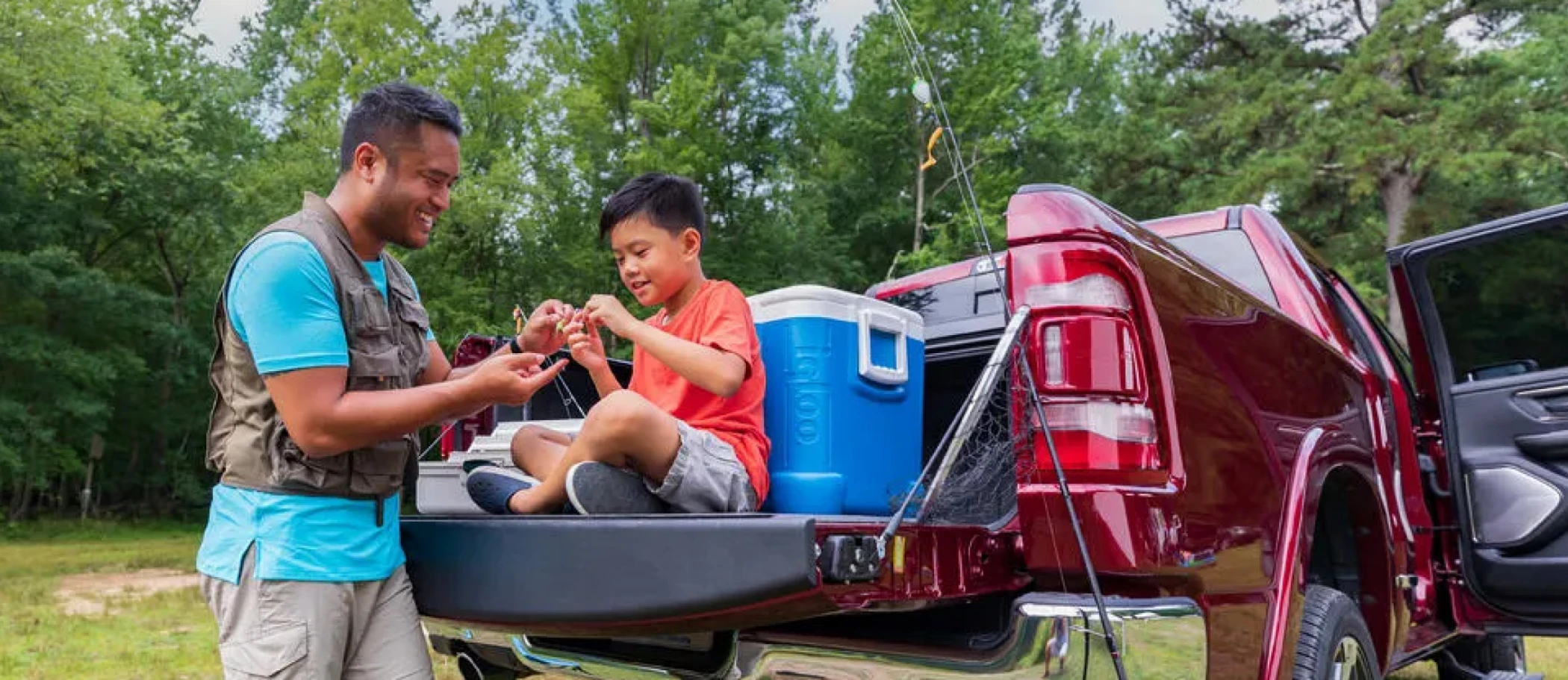 Boy is sitting on a truck tailgate while his smiling father helps him set up his fishing rod