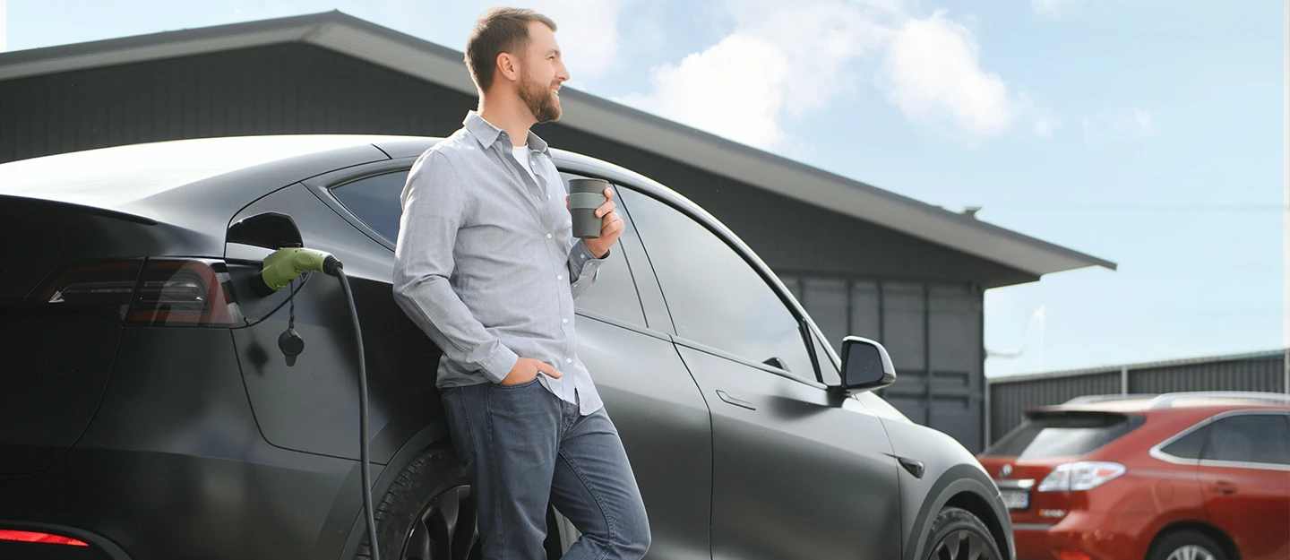 man leaning on his tesla as it charges while sipping coffee