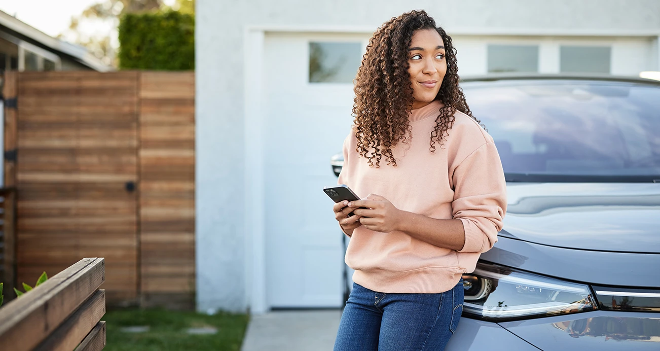 A woman casually leaning against a car in her driveway, her phone in hand as she researches information about buying a car 