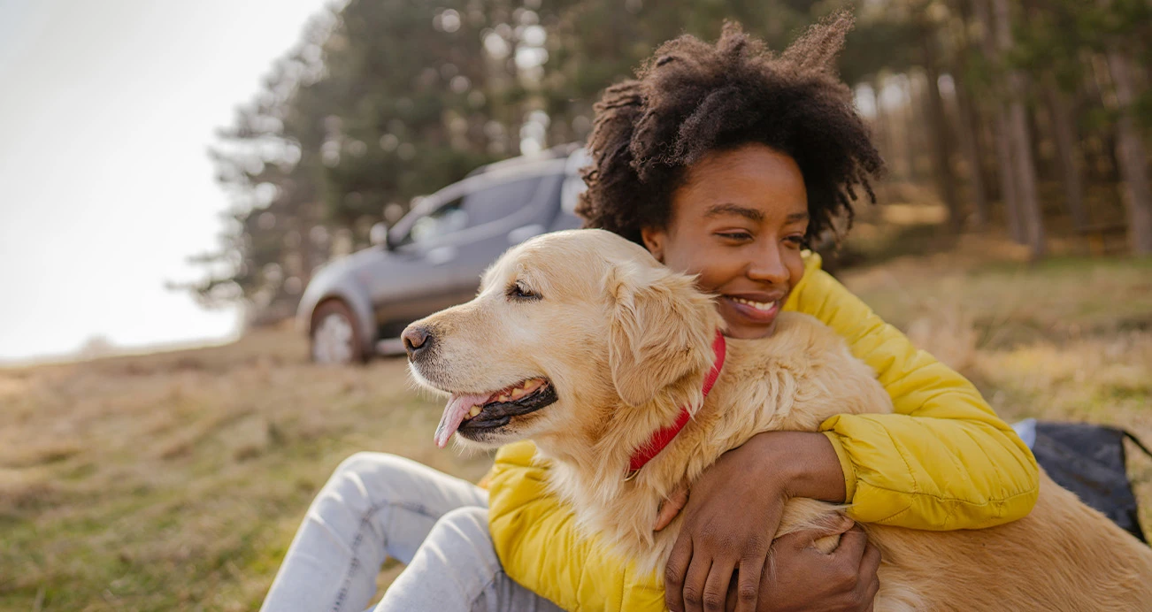 Woman sitting on grass smiling while hugging golden retriever. Car and trees in background