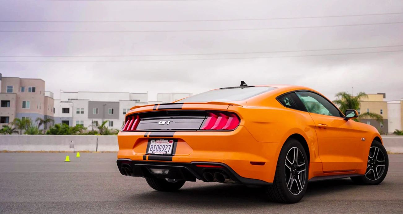 Rear shot of an orange Mustang with a black stripe parked in a parking lot with buildings in the background