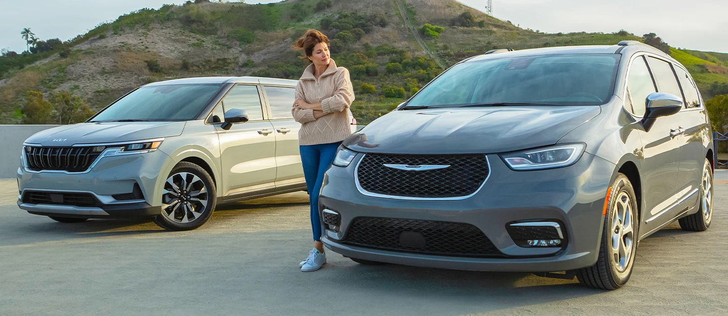 Host stands between 2024 Chrysler Pacifica and Kia Carnival arms crossed comparing the two
