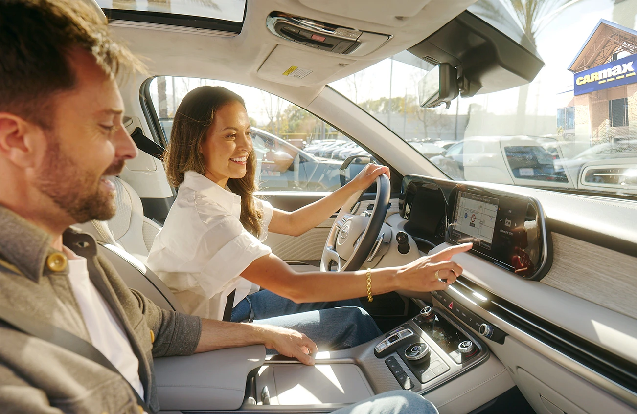 Woman in drivers seat with passenger they are smiling as they look at the infotainment system in a car on the CarMax store lot