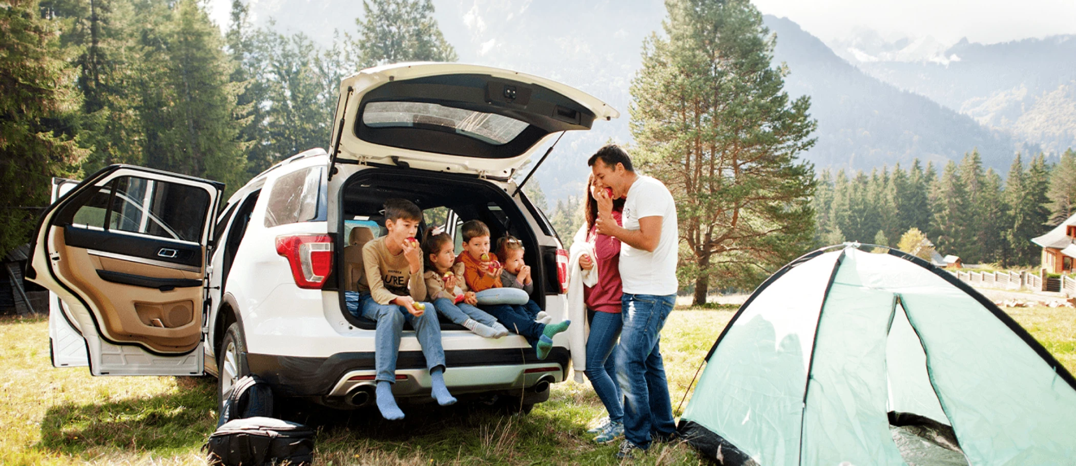 Family sitting in open trunk of white SUV next to camping site