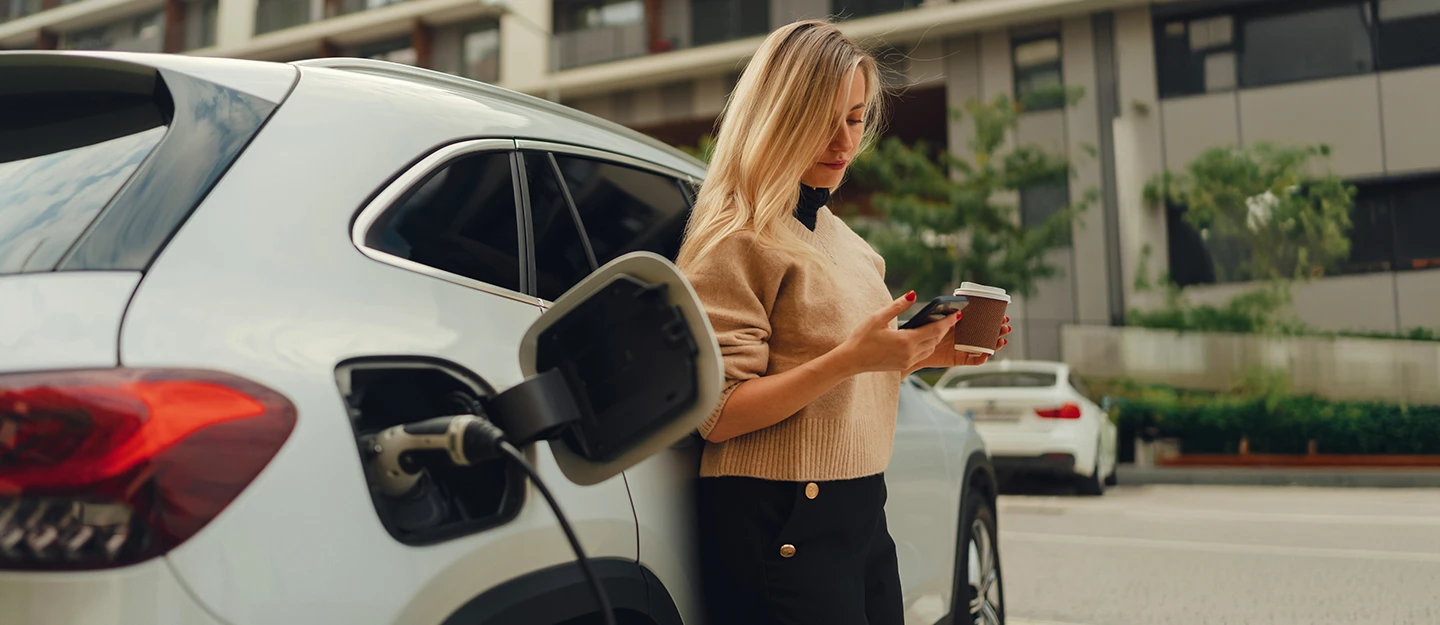 Woman leans against white EV while it is charging. She holds a phone and coffee cup in her hands.